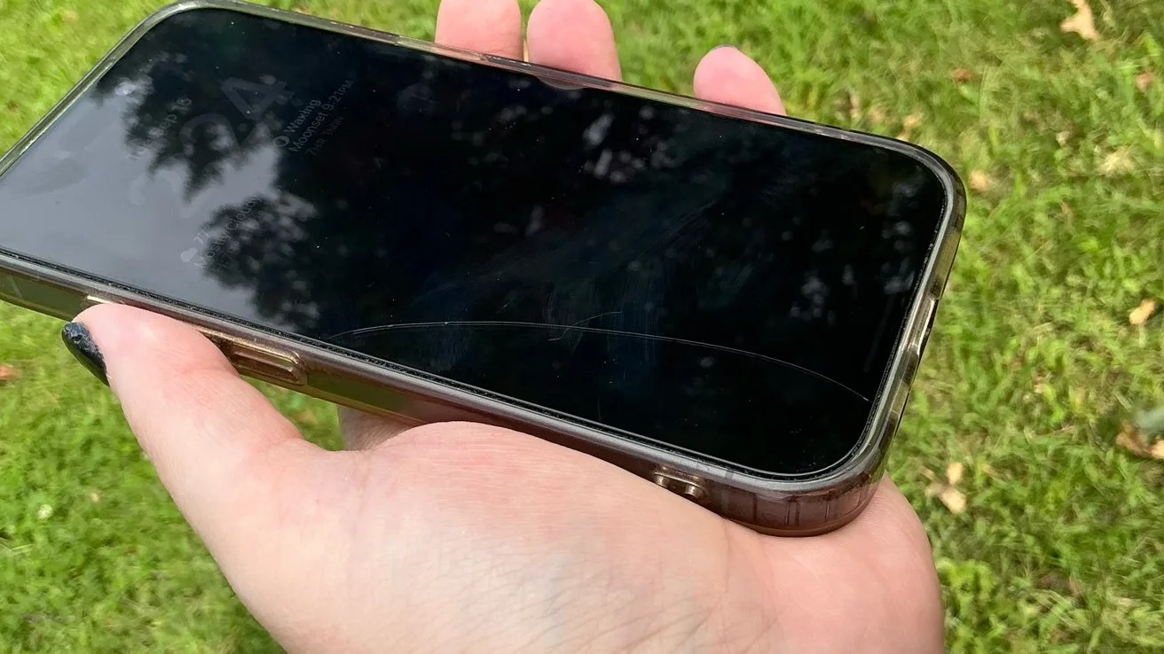 Hand holding a smartphone with a dark screen showing reflections of trees and a noticeable crack across the lower part, against a bright green grassy background