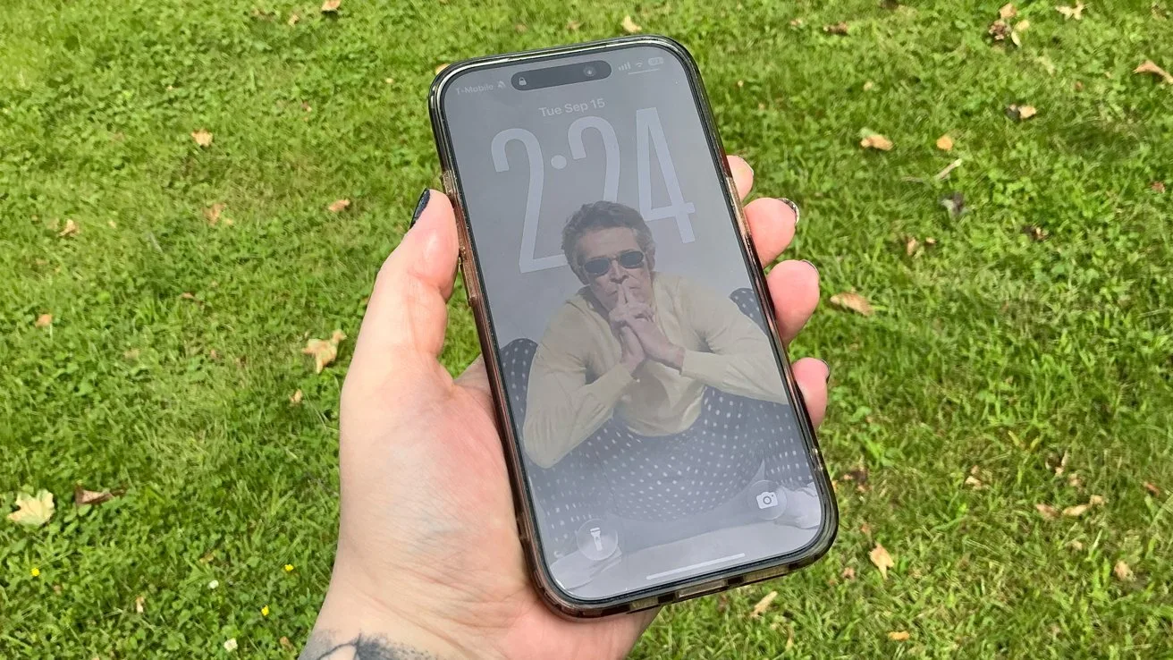 Hand holding a smartphone outdoors over green grass, screen showing lock screen with faint reflection and a person wearing sunglasses and light-colored clothes as the wallpaper