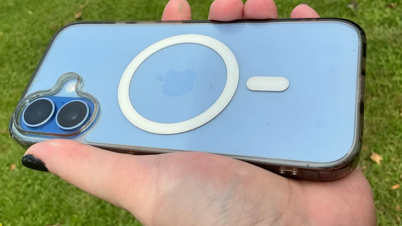 Hand holding a light blue iPhone with MagSafe circle and pill symbol on the back, dual camera lenses in a clear case, against a grassy outdoor background