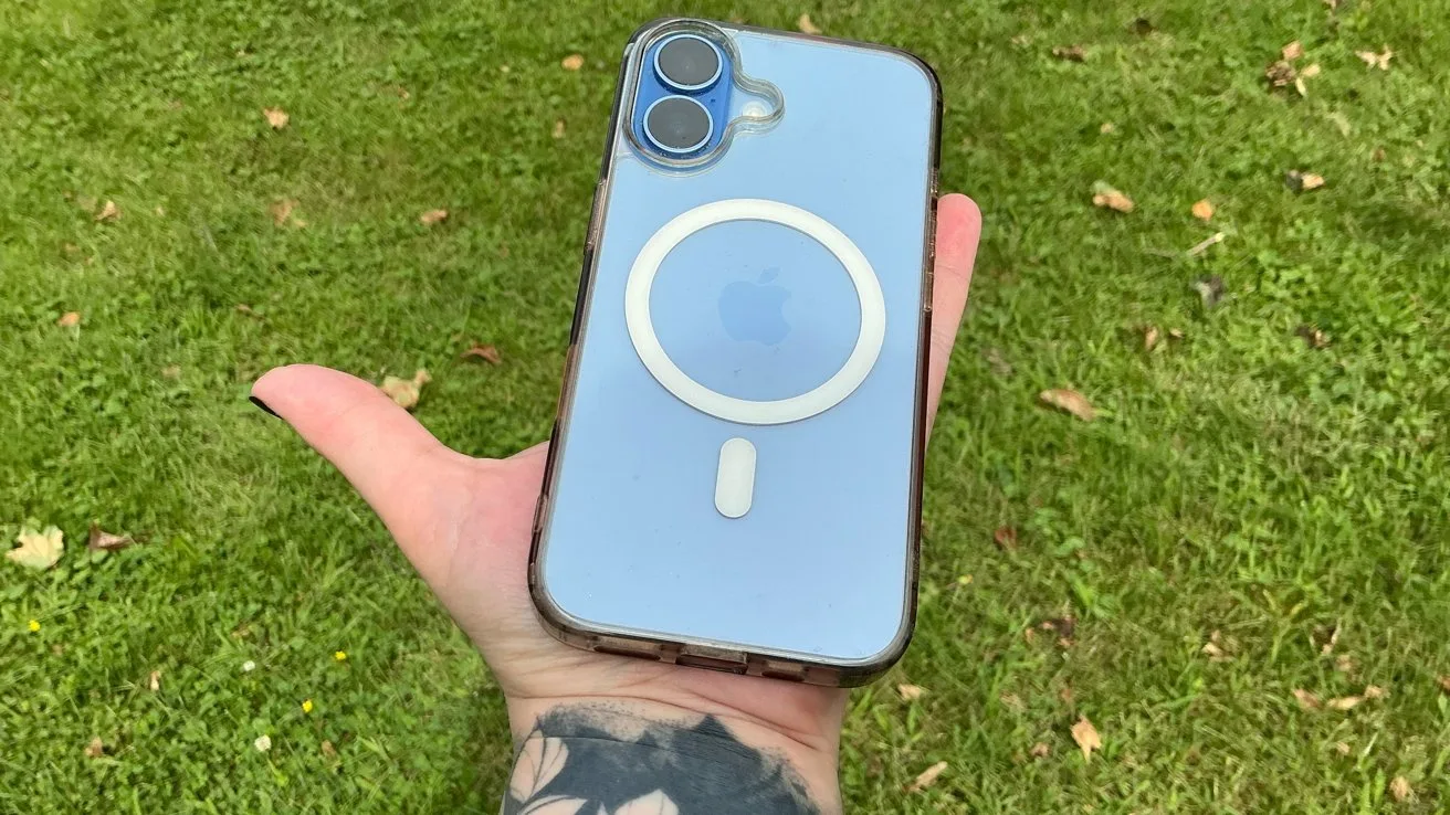 Tattooed hand holding a blue iPhone with clear MagSafe case and dual camera lenses, centered against a bright green grassy background with a few scattered leaves