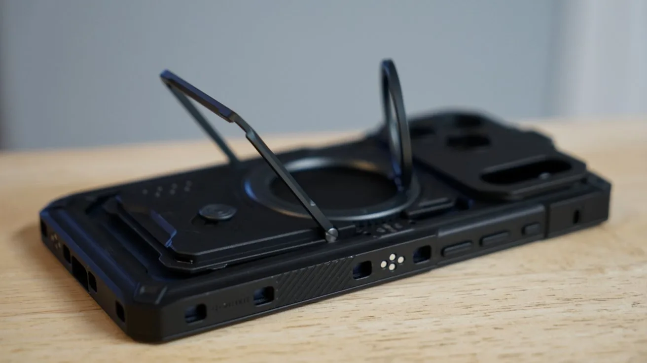 Black rugged iPhone case lying on a wooden surface, featuring a fold-out metal kickstand and circular ring stand, with cutouts for buttons, ports, and rear camera module