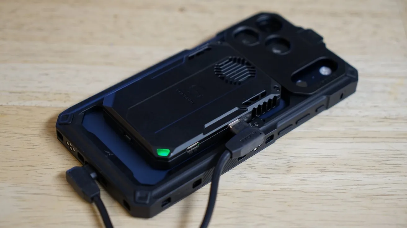iPhone lying face down on a wooden surface, with a black external drive magnetically attached to its back and connected by a short USB cable