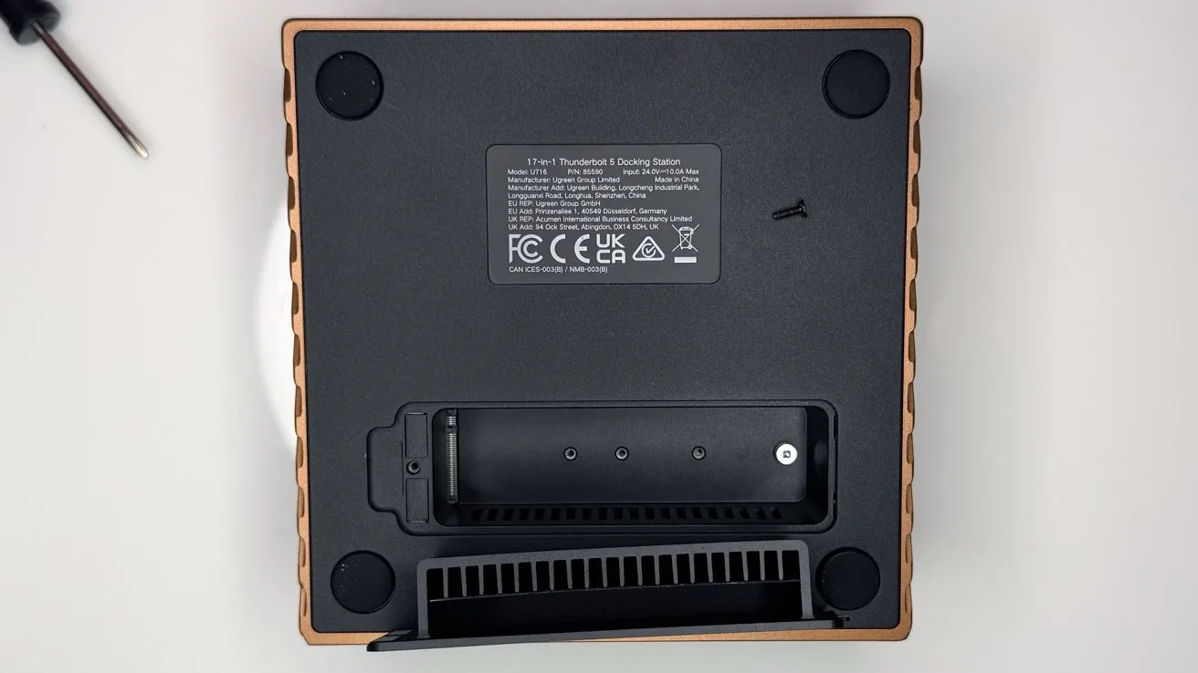 Bottom view of a square electronic device with four rubber feet, central label with certification logos, a removable battery compartment, ventilation grill, and a screwdriver partially visible on the side