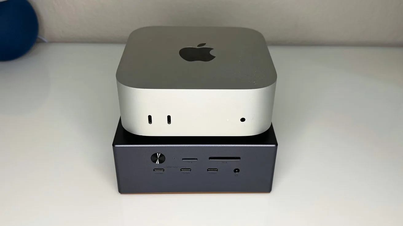 Small silver Apple desktop computer stacked on top of a darker rectangular mini PC, both resting on a glossy white surface against a plain light-colored wall.
