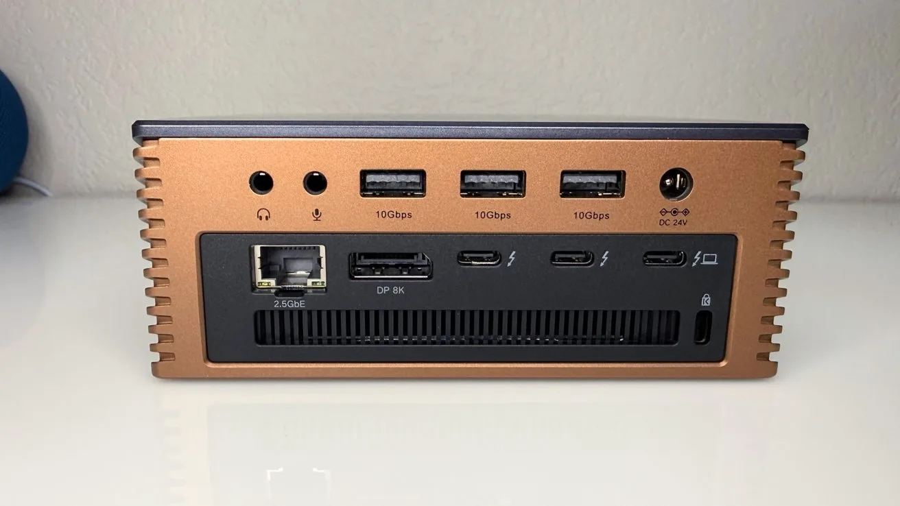 Compact copper-colored mini PC rear panel showing audio jacks, three USB ports, power connector, Ethernet port, three USBC or DisplayPort ports, vent grille, and status light on white desk