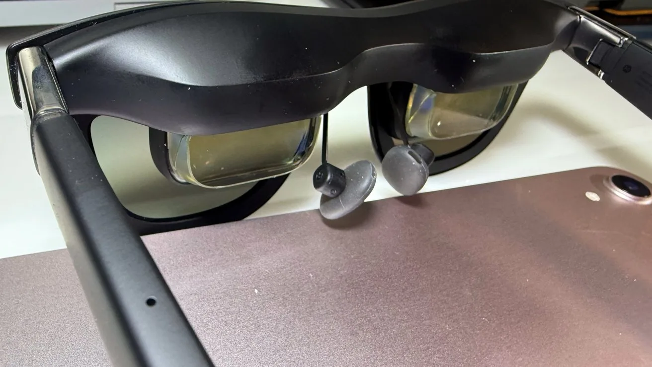 Black augmented reality glasses on a surface, with attached earbuds visible, alongside a metallic tablet or device in the background.