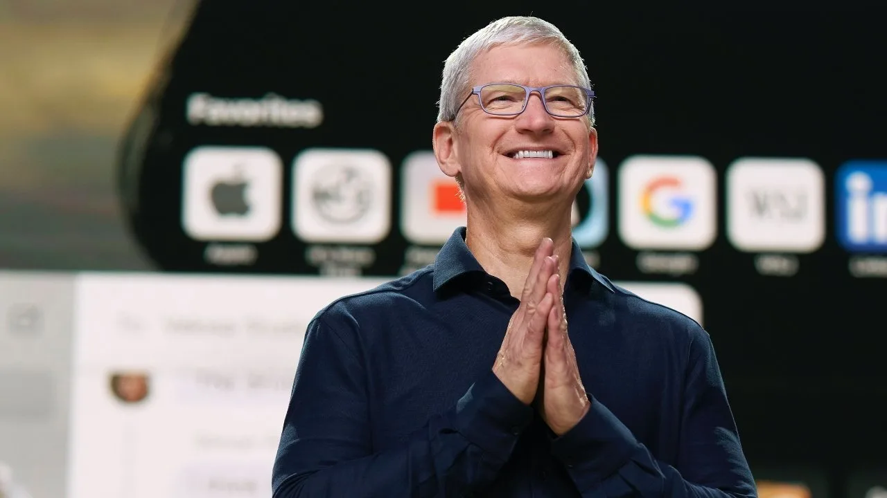 Smiling man with gray hair and glasses stands on a stage, hands clasped, in front of a large screen displaying various app icons and a favorites toolbar