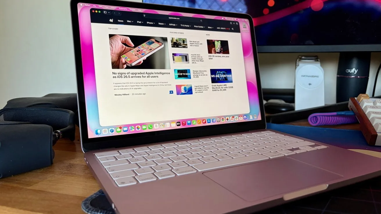 Open pink laptop on a desk displaying a news website, with a larger monitor, external keyboard, and various office accessories in the background