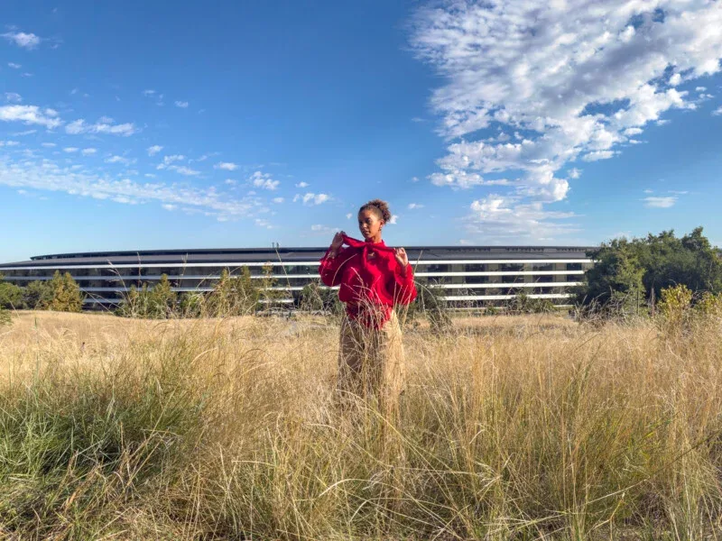 A person with curly hair wearing a red blouse and tan pants stands in a field of tall, dry grass in front of a long, modern building under a blue sky with scattered clouds.