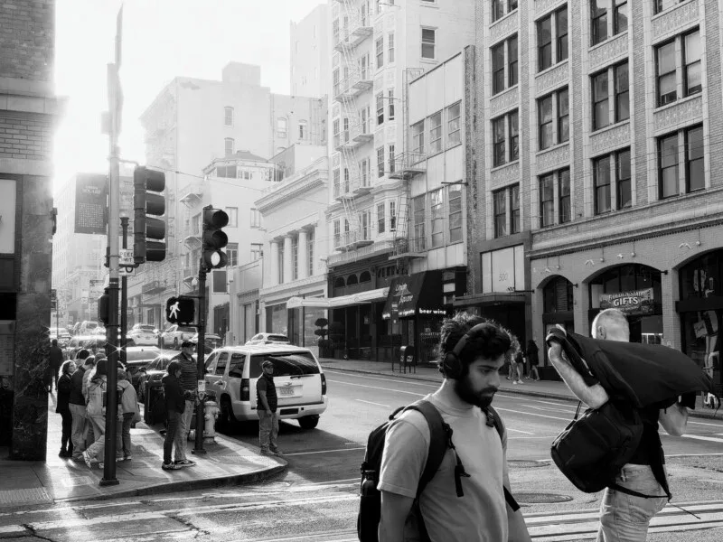Pedestrians wait at a street corner in a city while a man with a backpack and headphones walks across the foreground.