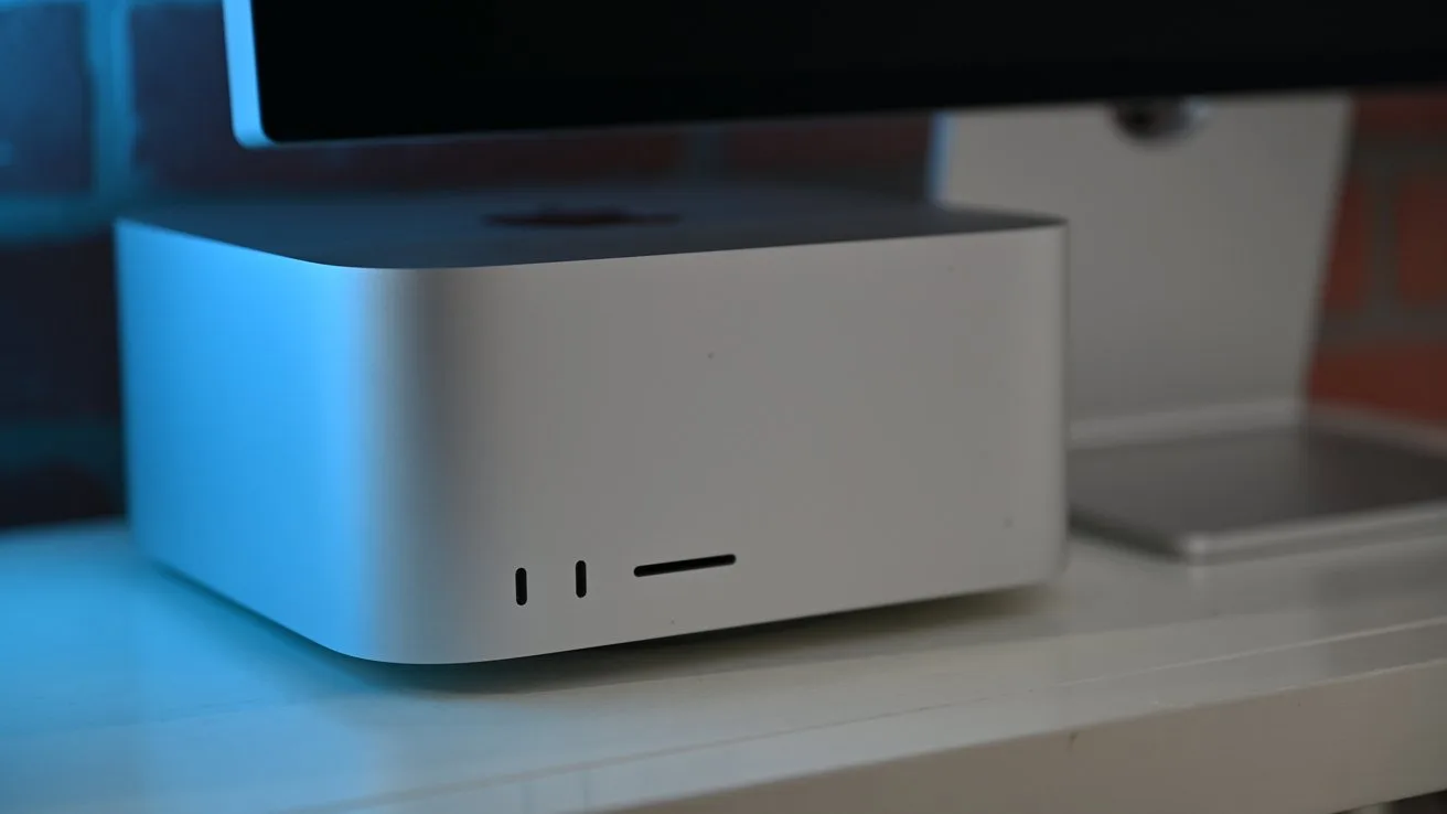 Mac Studio on a white desk beneath a large monitor, softly lit with blue light, showing front ports and minimalistic modern design