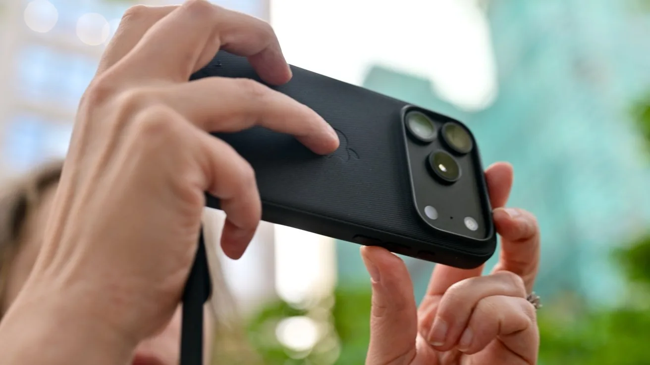 Hands holding a black iPhone 18 Pro in a case horizontally, focusing on its large dual rear cameras, with blurred city buildings and greenery in the background