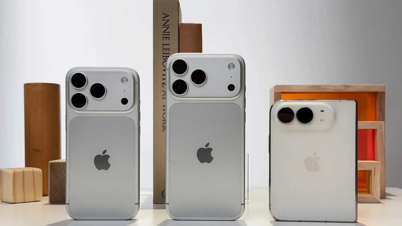 Three silver iPhones standing upright on a table, showing backs with large camera lenses, arranged by height against a soft, minimalist background with books and wooden decorative blocks