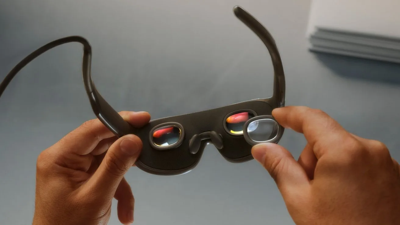Hands holding Meta VR Glasses, showing curved dark frame and reflective lenses with small colorful displays, on a smooth gray tabletop with a blurred stack of papers in the background