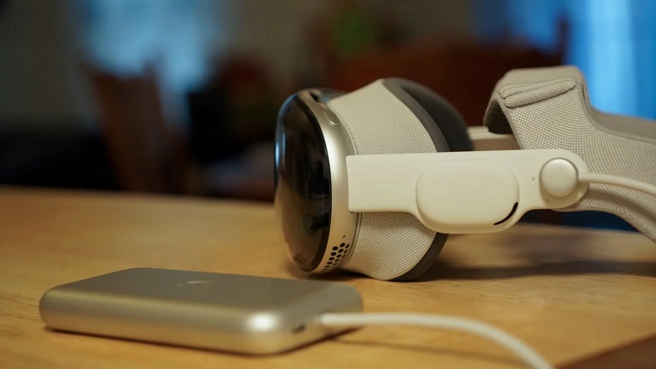 Apple Vision Pro resting on a wooden table beside a slim rectangular device connected by a cable, in a softly lit indoor setting