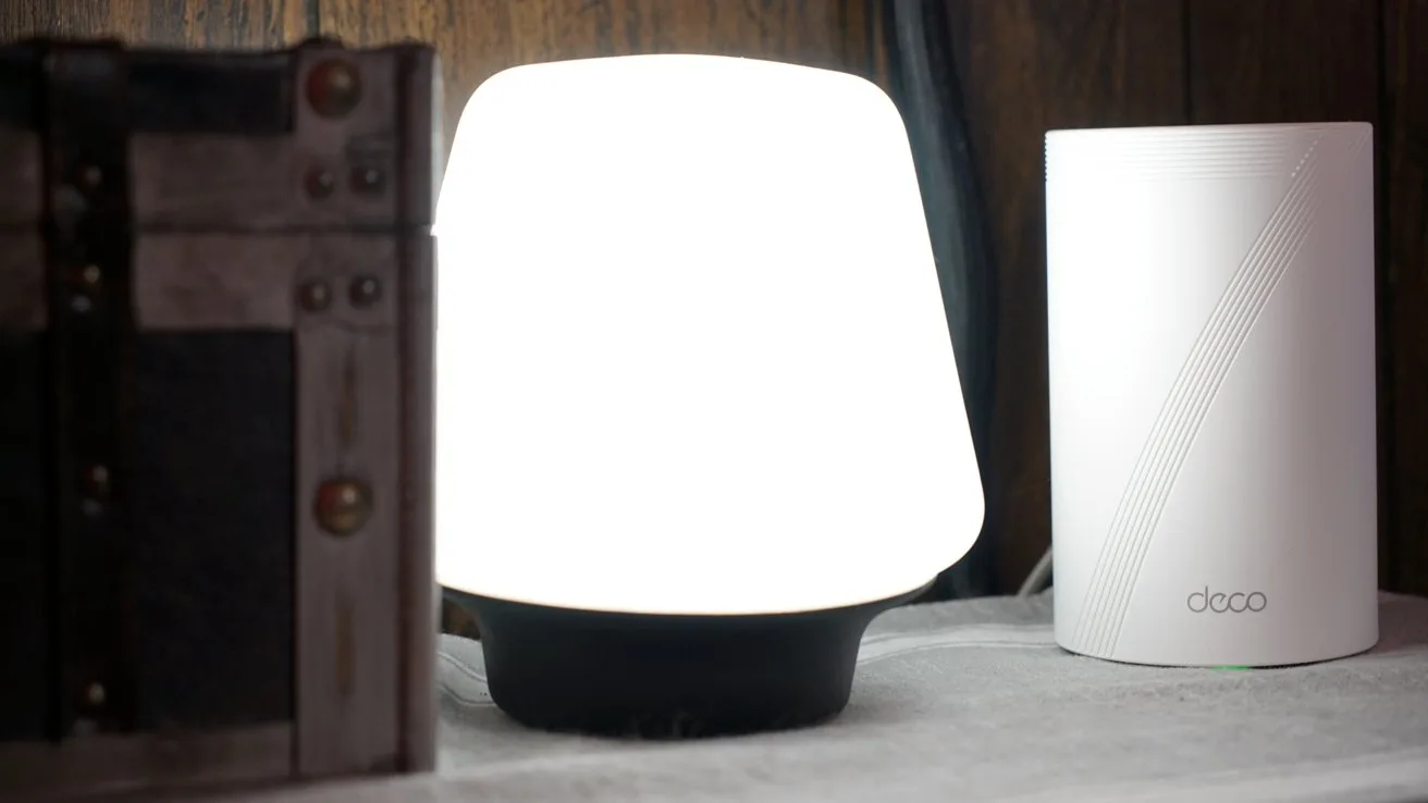 Warmly lit modern lamp beside a white cylindrical device with subtle line design on a wooden surface.
