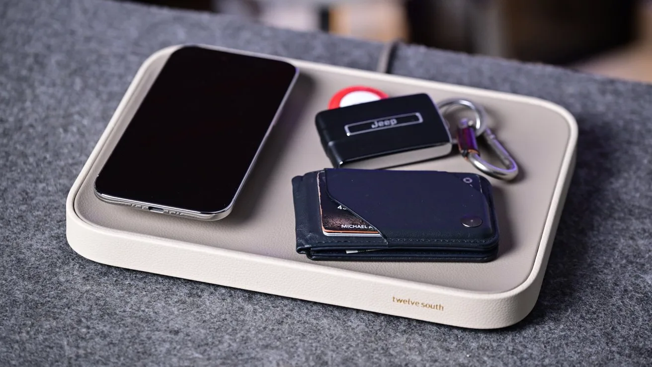 Beige desk tray holding a smartphone, slim wallet with cards, car key fob, metal keyring, and small round tracker, resting on a gray fabric surface
