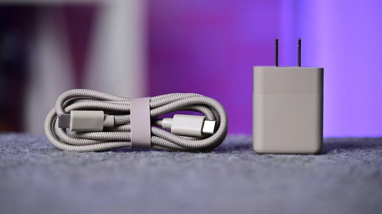 Coiled gray USB-C charging cable beside a matching rectangular wall power adapter, both resting on a soft gray surface with a blurred purple background