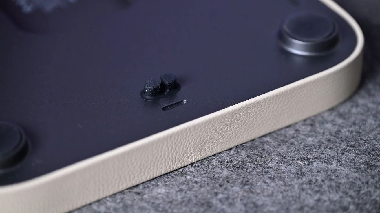 Close-up of a device's rounded corner showing textured beige edge, dark underside with two rubber feet, a small switch, and a narrow slot, resting on gray fabric surface