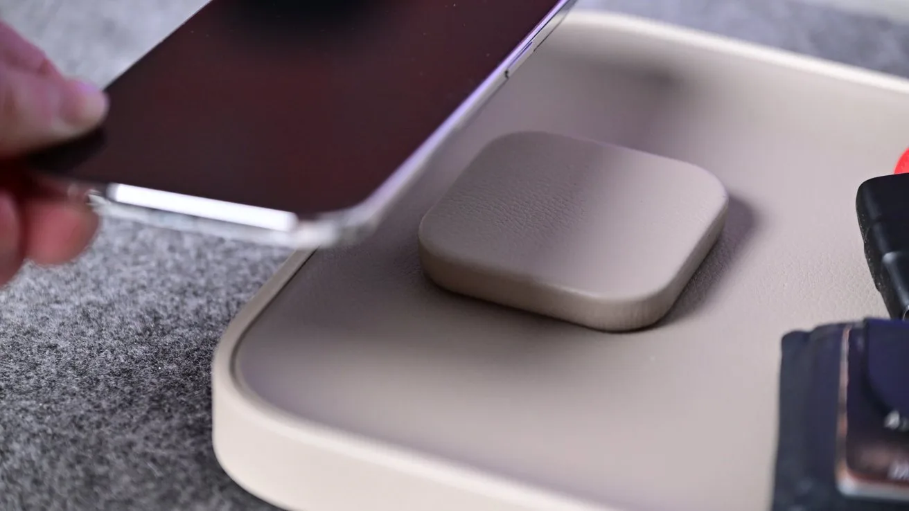 Hand holding a smartphone above a small square wireless charging pad on a tray, with keys and a wallet nearby on a textured surface