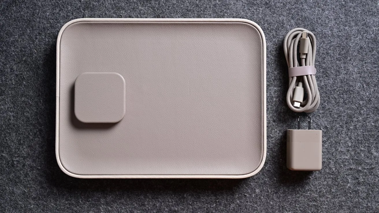 Minimalist beige wireless charging pad with a raised square section on the left, placed beside a neatly coiled USB cable and a small matching power adapter on dark fabric background