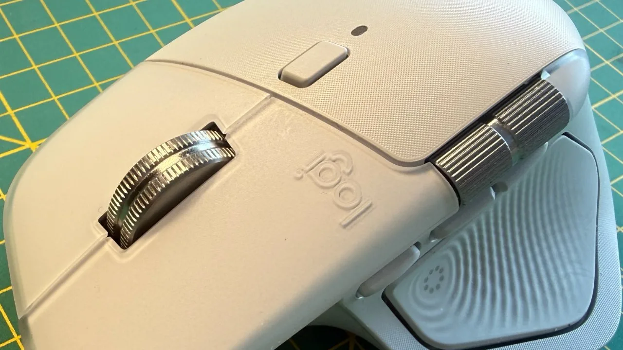 Ergonomic white computer mouse with metallic scroll wheel and side dial, resting on a green and yellow grid-patterned surface.
