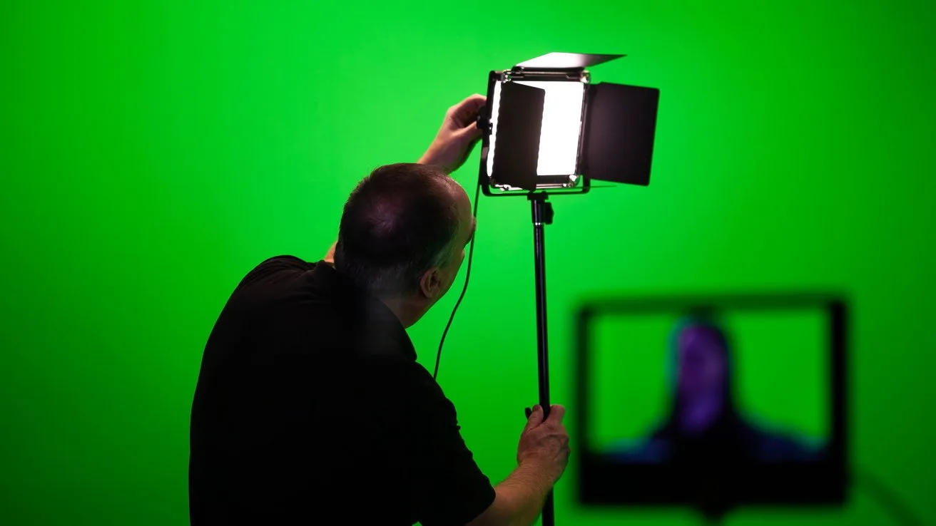 Person adjusting a bright studio light against a solid green screen background, with a blurred monitor in the foreground showing an indistinct figure
