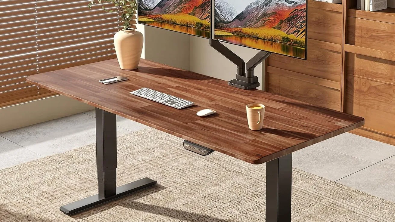 Wooden standing desk with black legs, holding dual monitors on a mount, keyboard, mouse, coffee mug, and vase, in a bright modern room with blinds and a rug