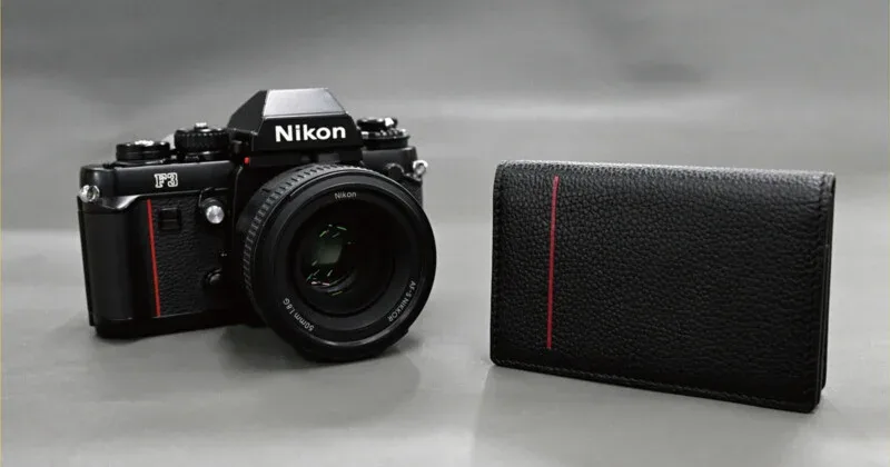 A black Nikon F3 film camera with a lens sits next to a black leather wallet featuring a vertical red stripe on a gray background.