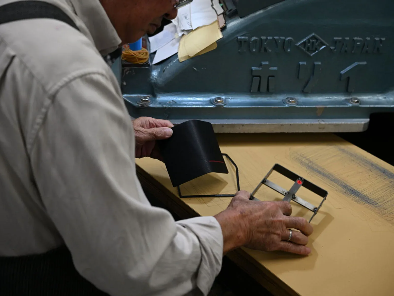 An elderly man in a light-colored shirt works with a leather piece and a metal die on a workbench in front of a Tokyo, Japan machine.