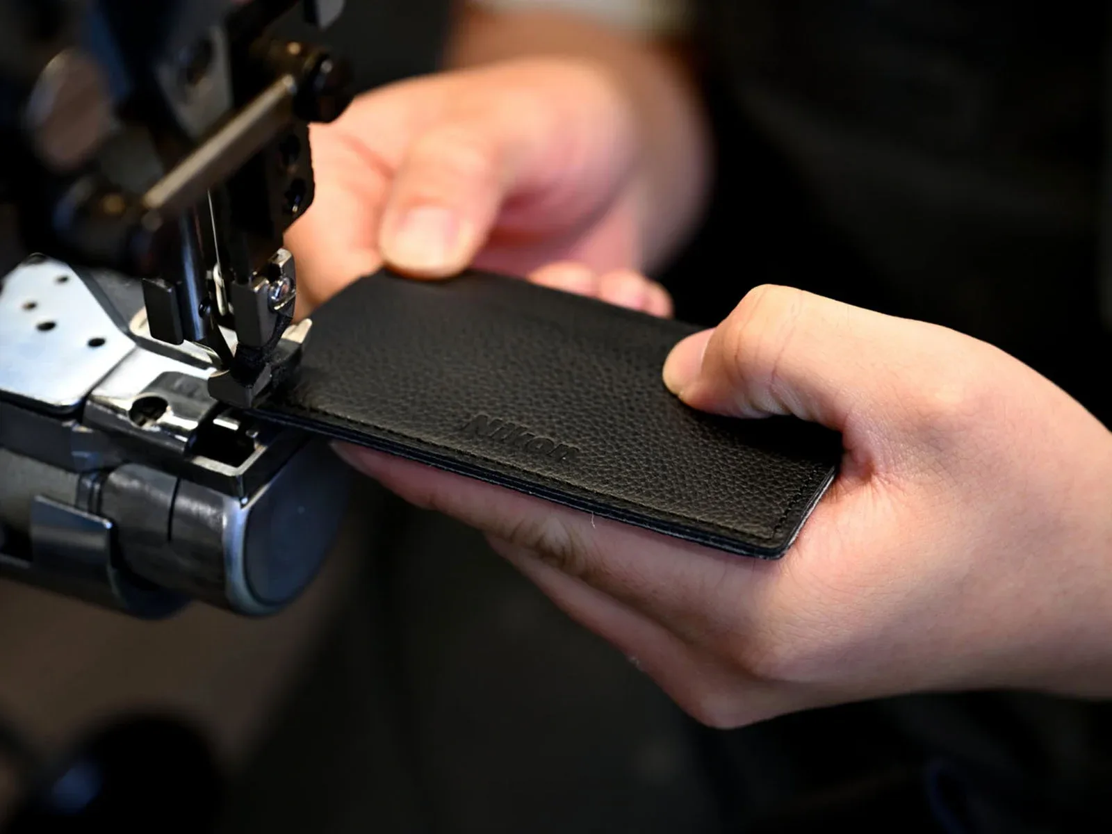 A person uses a sewing machine to stitch a piece of black textured leather embossed with the Nikon logo.