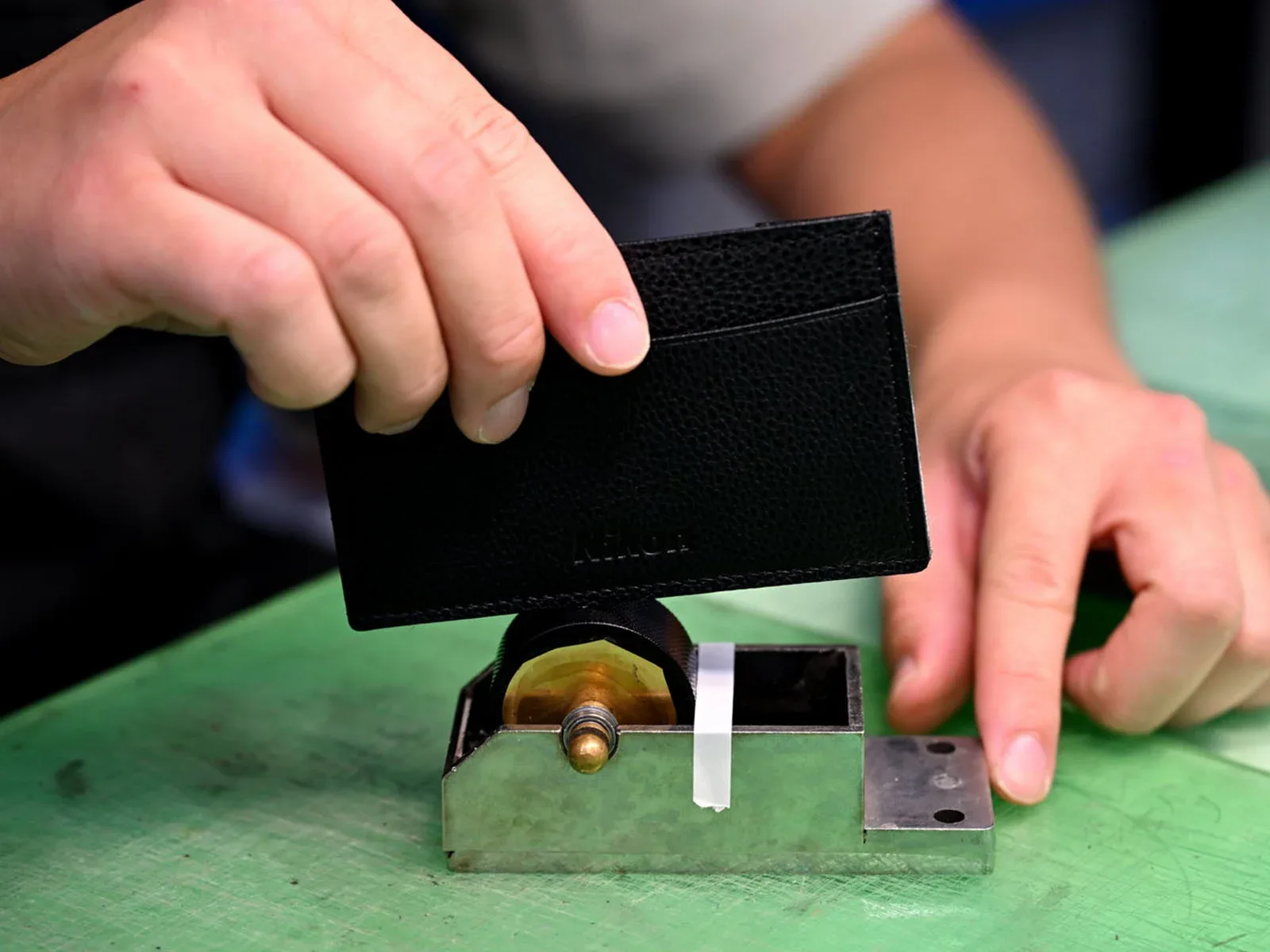 A person's hands press a black leather card holder with a debossed "Nikon" logo onto a metal stamping tool on a green surface.