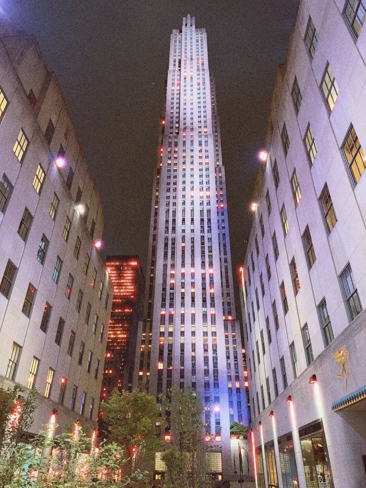 Tall illuminated skyscraper at night framed by two shorter office buildings, colorful lights reflecting on windows, with a small courtyard of trees and plants in the foreground