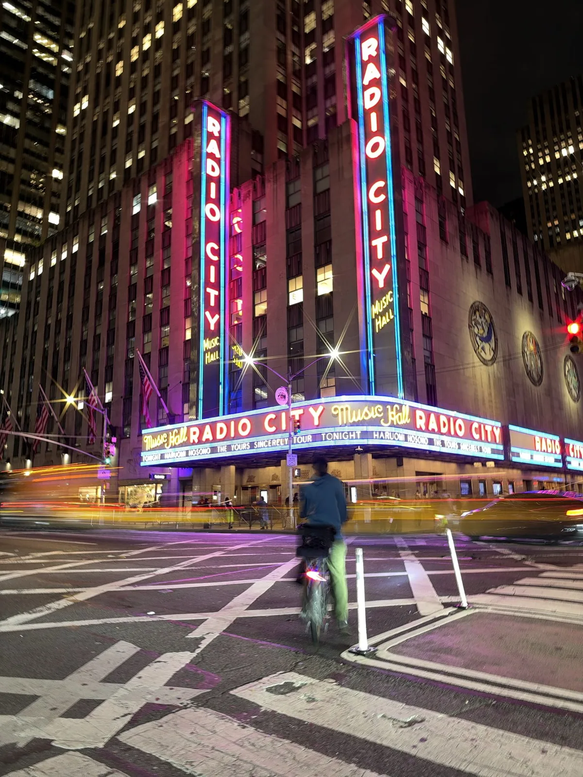 Nighttime street scene at Radio City Music Hall, neon signs glowing pink and blue, tall surrounding buildings, blurred car lights, and a cyclist crossing the wide striped intersection