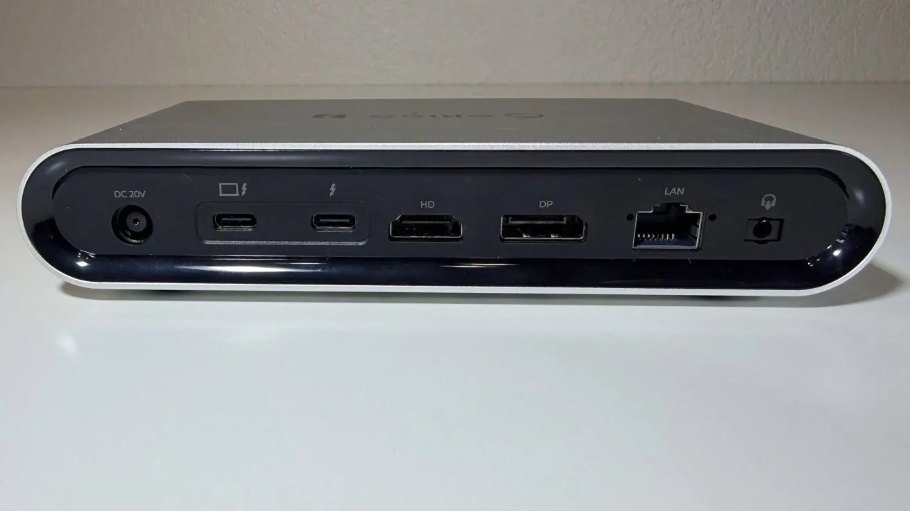 Small rectangular computer docking station on a desk, showing rear ports including power, USB-C, DisplayPort, HDMI, USB-A, Ethernet, and audio jack, with a smooth gray and black finish