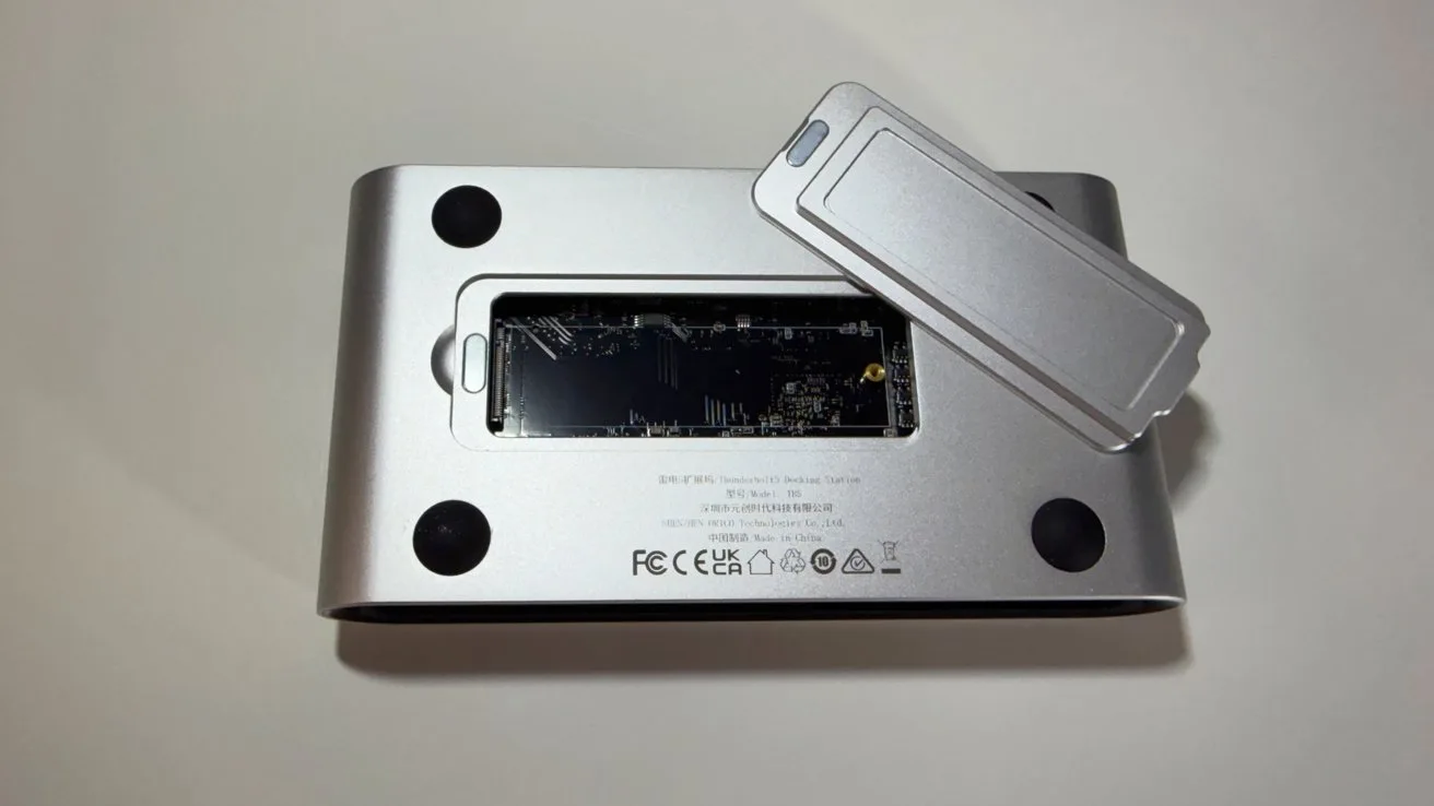 Silver electronic device flipped upside down with rectangular access panel removed, exposing internal circuit board, surrounded by four black rubber feet and certification markings on a plain light background