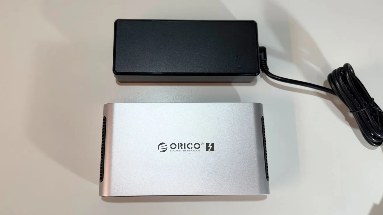 Silver ORICO external hard drive enclosure lying below a rectangular black power adapter with attached cable, both placed on a clean white surface