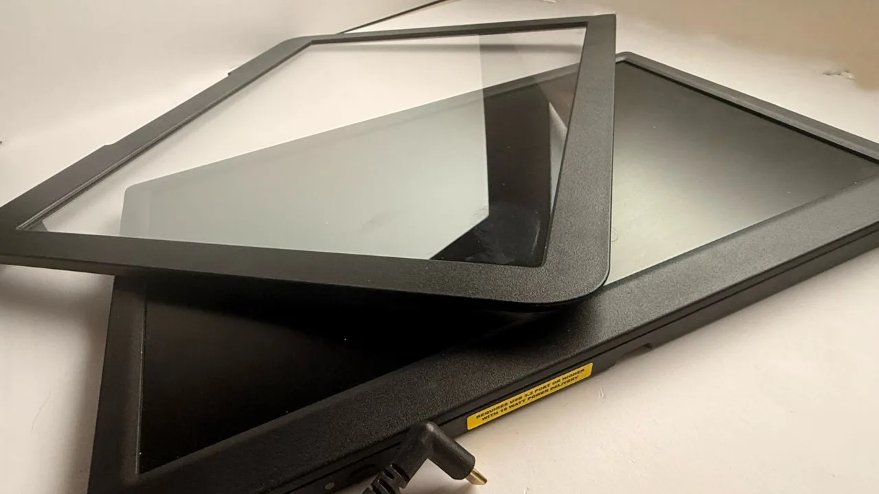 Two black electronic display screens stacked on a white surface, one with a clear protective glass frame resting diagonally on top, with a visible power connector cable in the foreground