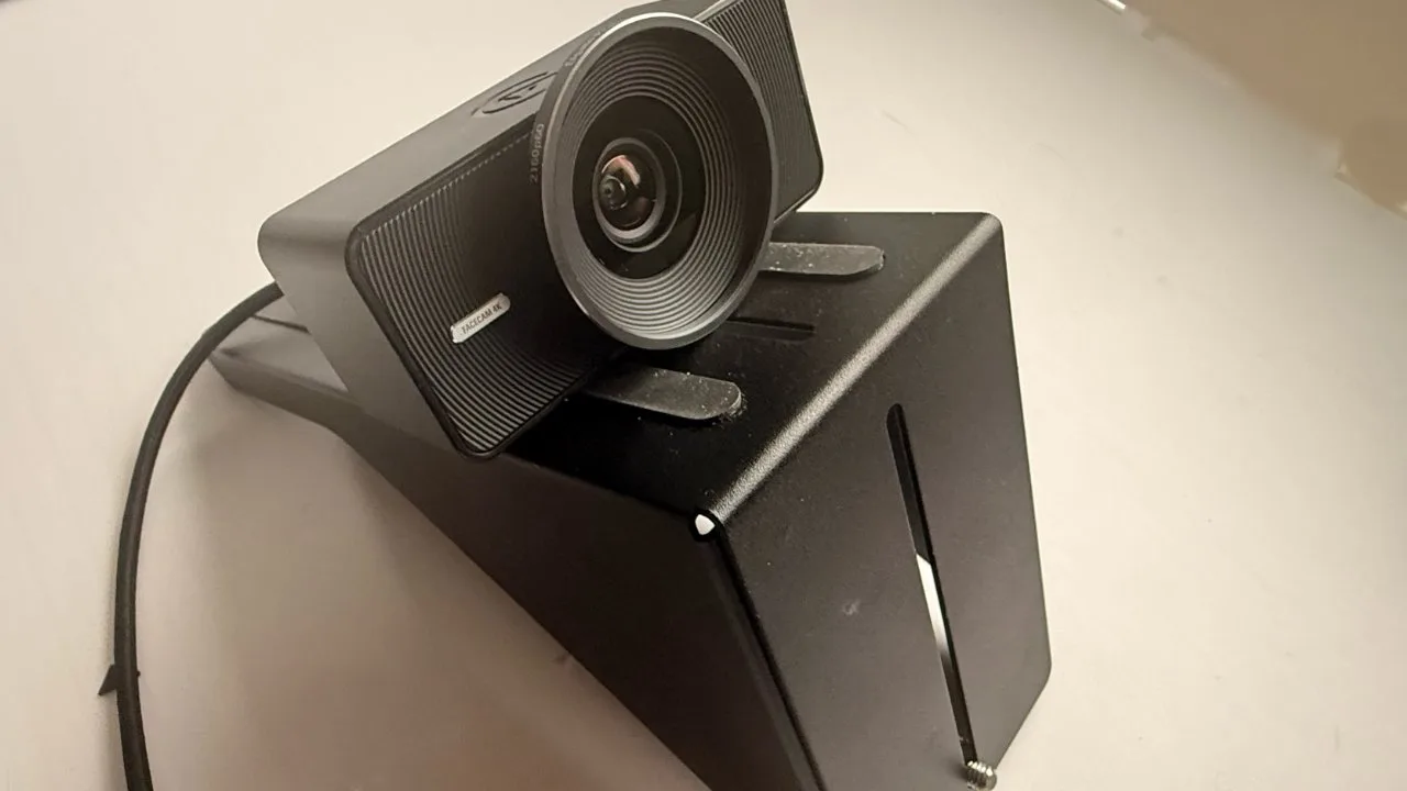 Black widescreen webcam with large circular lens mounted on an angled black metal stand, connected by a cable, sitting on a plain light-colored surface.