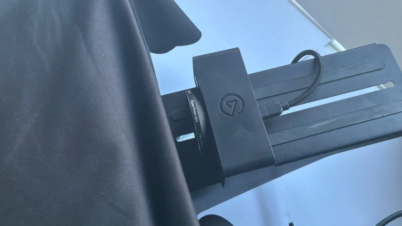 Close-up of an Elgato camera or capture device mounted on a black adjustable bracket, partly covered by a dark fabric backdrop on the left, against a light background.