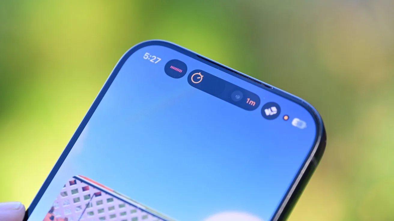 Close-up of a smartphone's top screen showing front camera cutout, status icons, and a timer, with a clear blue sky and blurred green background behind the phone