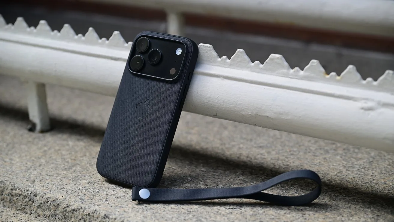 Black smartphone with triple rear cameras in a dark case, propped against a white metal railing on concrete, with a matching black wrist strap lying beside it