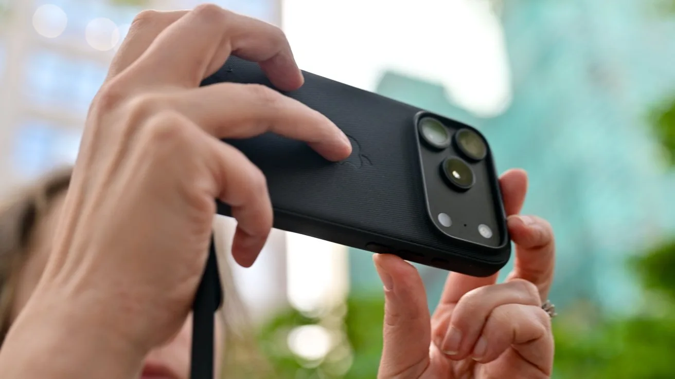 Person holding a smartphone horizontally to take a photo outdoors, focusing on the phone's dual camera lenses and protective case, with blurred buildings and greenery in the background
