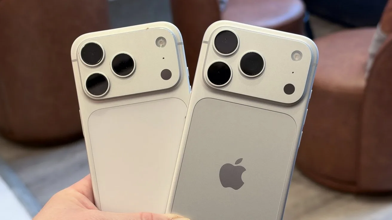 Hand holding two new iPhones with large dual rear cameras, one white and one light gray, showing the Apple logo and rounded rectangular camera bump in a casual indoor setting