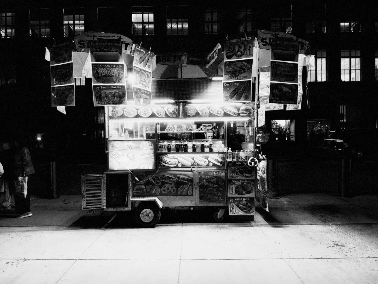 Brightly lit street food cart at night on a city sidewalk, surrounded by hanging menus and signs, standing alone against a dark building background in black and white photography