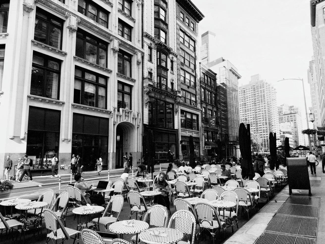 Outdoor cafe tables and chairs line a busy city sidewalk beside tall historic buildings, with people walking and dining in a lively urban street scene, shown in black and white