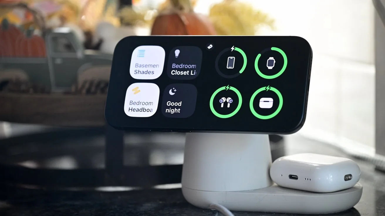 Smartphone on a white stand displaying home automation controls with circular green icons, next to a small white charging case on a countertop in a softly lit room