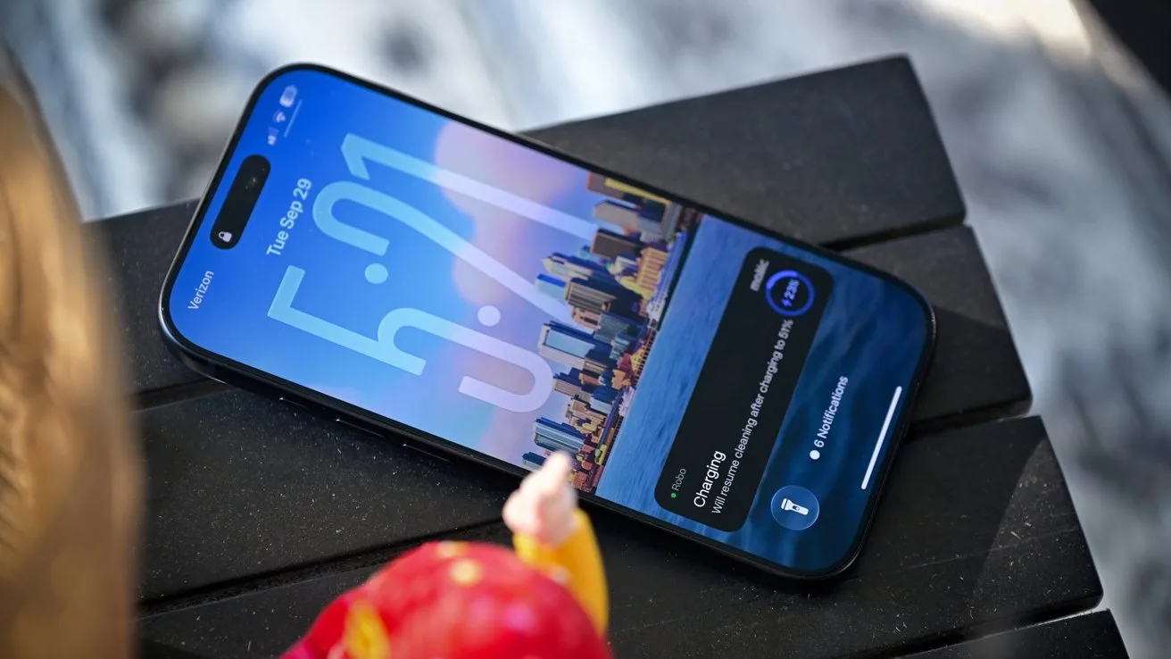 Smartphone lying on a dark slatted surface, screen on, showing a cityscape lock screen with large digital clock and notifications, with a colorful blurred foreground object near the bottom edge