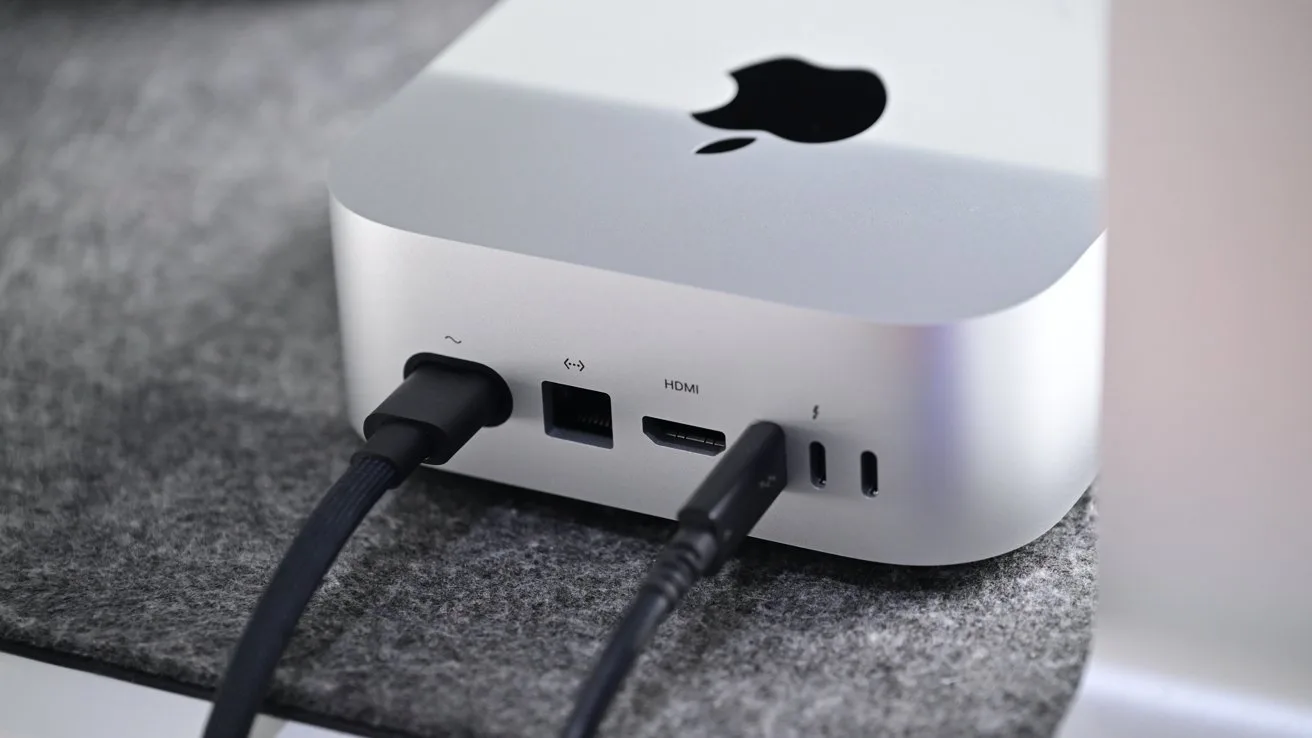Silver Apple Mac mini on a gray desk surface, shown from the back with power, HDMI, USB, and audio cables plugged into its various ports