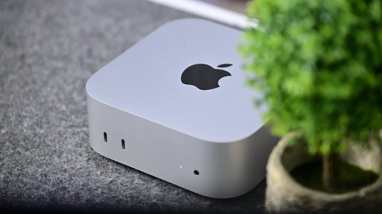 Small silver Apple Mac mini desktop computer on a gray surface, featuring the Apple logo on top, with a blurred green potted plant in the foreground